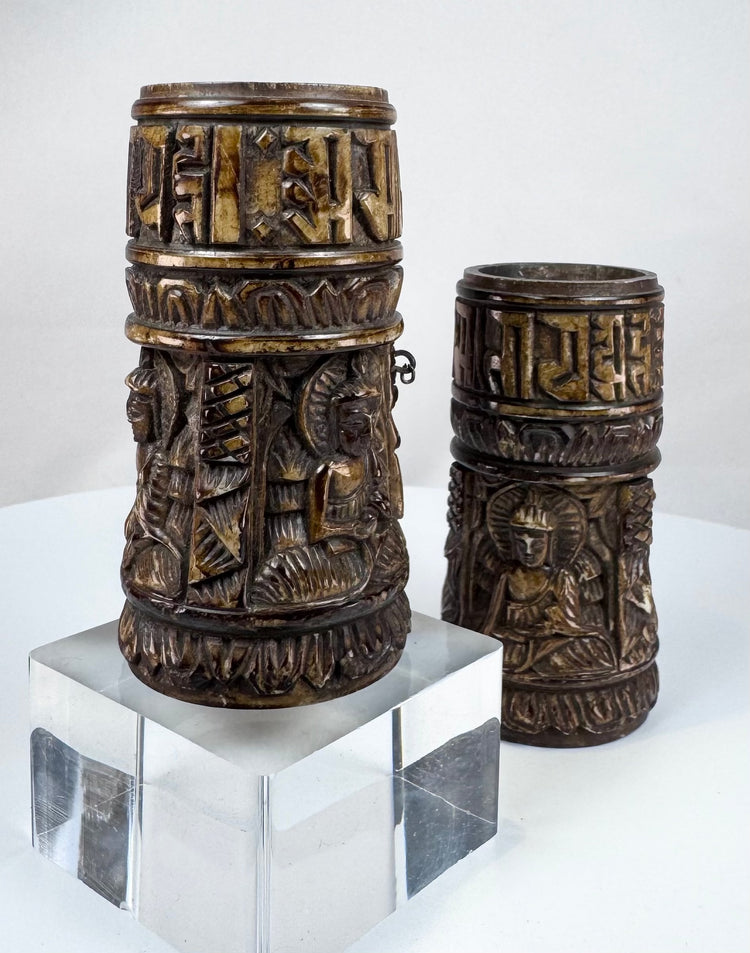 Detailed carving on a Tibetan Buddhist prayer wheel showing relief Buddha figures beneath a band of sacred inscriptions.