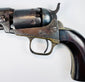 Close-up of the Colt Model 1849 Pocket Revolver’s worn cylinder engraving, mottled steel frame, and curved brass trigger guard.