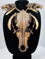 Skull and tooth necklace with layered cord bindings, dark bone cavities, and mottled golden-brown surfaces on a black bust.