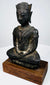 Crowned bronze Buddha with textured metal surfaces and an oval bronze seat mounted on a rectangular wooden base.
