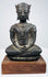 Crowned bronze Buddha seated with crossed legs and folded hands, mounted on a wooden base against a beige background.