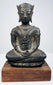 Crowned bronze Buddha seated with crossed legs and folded hands, mounted on a wooden base against a beige background.