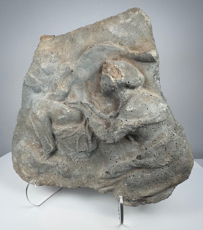 Roman relief fragment in gray volcanic tuff showing weathered human figures and naturally pitted stone surface