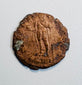 Late Roman copper-alloy coin with standing figure reverse, worn inscription, edge losses, and areas of green oxidation.