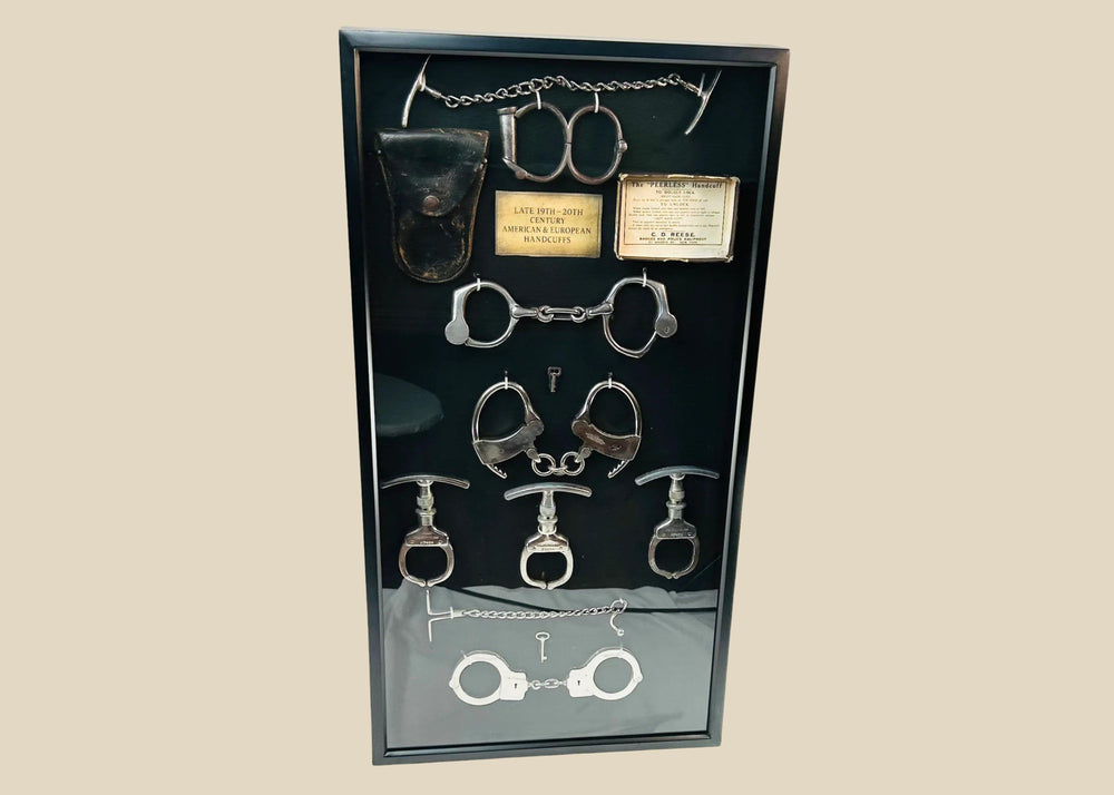 Framed shadow box display featuring a chronological collection of American and European handcuffs, leg irons, mechanical nippers, chain restraints, keys, and original leather pouches, mounted on black fabric with synthetic plaque, showcasing restraint devices from circa 1880s to 1950s.