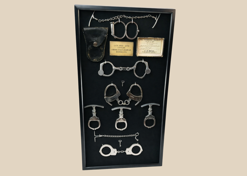 Framed shadow box display featuring a chronological collection of American and European handcuffs, leg irons, mechanical nippers, chain restraints, keys, and original leather pouches, mounted on black fabric with synthetic plaque, showcasing restraint devices from circa 1880s to 1950s.