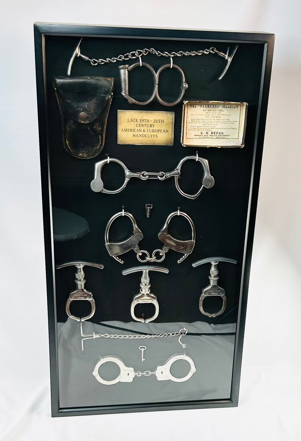Framed shadow box display featuring a chronological collection of American and European handcuffs, leg irons, mechanical nippers, chain restraints, keys, and original leather pouches, mounted on black fabric with synthetic plaque, showcasing restraint devices from circa 1880s to 1950s.