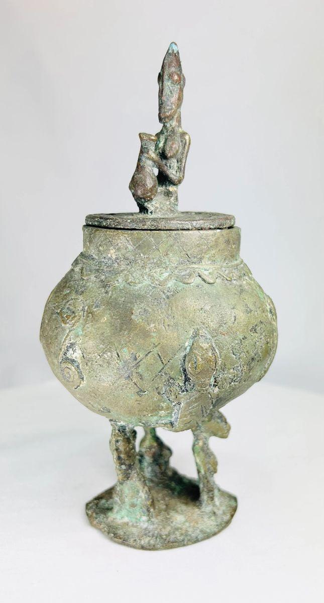 19th-century Dogon bronze container from Mali with bulbous body, engraved motifs, anthropomorphic legs, and a sculpted human figure on the lid, used in ritual and healing practices.