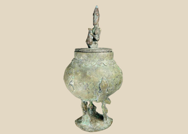 19th-century Dogon bronze container from Mali with bulbous body, engraved motifs, anthropomorphic legs, and a sculpted human figure on the lid, used in ritual and healing practices.
