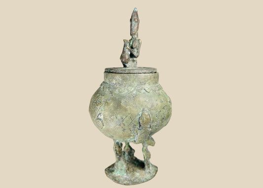 19th-century Dogon bronze container from Mali with bulbous body, engraved motifs, anthropomorphic legs, and a sculpted human figure on the lid, used in ritual and healing practices.