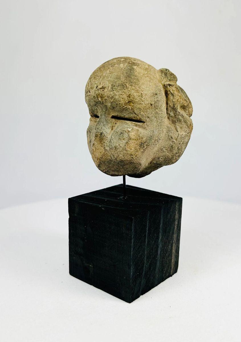 Ancient Mesoamerican stone mask fragment with minimalist facial features and narrow eye slits, carved from a single block of stone and mounted on a black wooden display stand, dating to 500 BCE–500 CE.