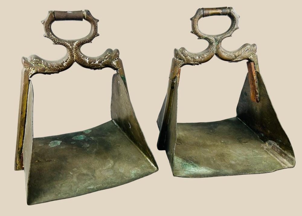 Antique pair of heavy brass equestrian stirrups with triangular footrests and intricately cast serpent motifs forming the handles, exhibiting detailed craftsmanship, natural patina, and symbolic protective design elements often associated with strength and guardianship in traditional horsemanship.