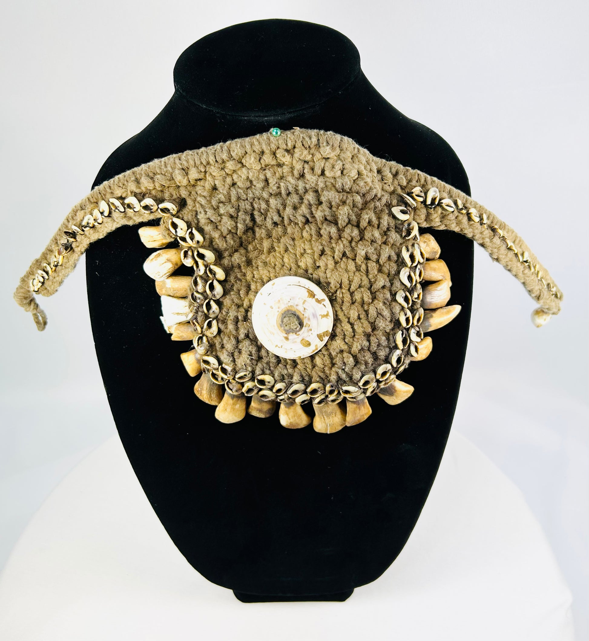 Antique ceremonial adornment from Papua New Guinea, late 19th–early 20th century, woven plant fiber with central cowrie shell and fringe of pierced animal teeth, displayed on a black stand.