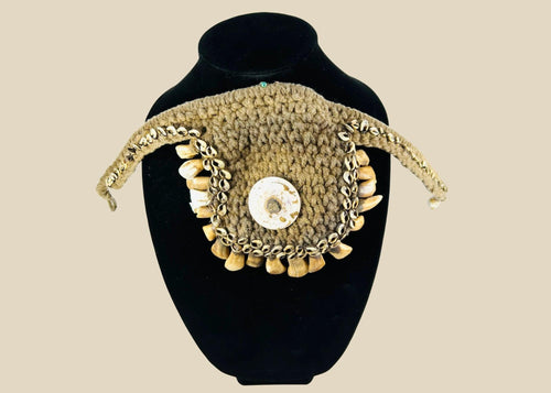 Antique ceremonial adornment from Papua New Guinea, late 19th–early 20th century, woven plant fiber with central cowrie shell and fringe of pierced animal teeth, displayed on a black stand.