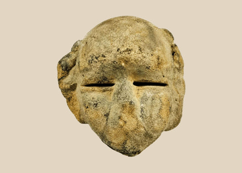 Ancient Mesoamerican stone mask fragment with minimalist facial features and narrow eye slits, carved from a single block of stone and mounted on a black wooden display stand, dating to 500 BCE–500 CE.