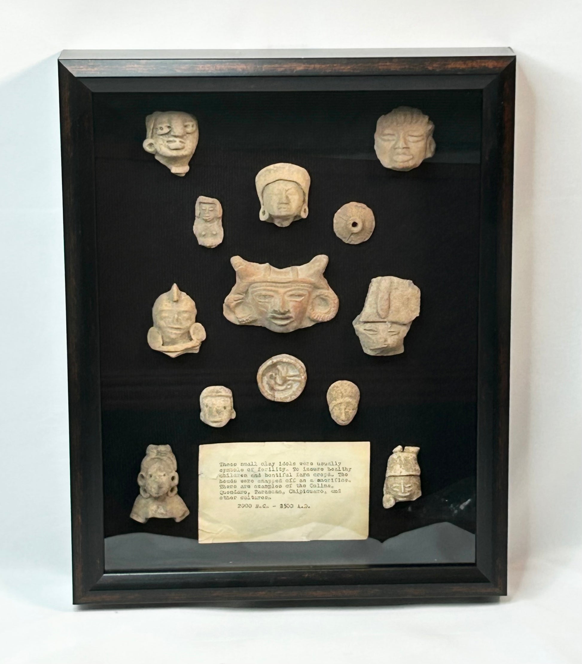 Shadowbox of 13 Pre-Columbian Mesoamerican terracotta fragments, including heads, maskettes, figurines, and spindle whorls from Teotihuacan and Veracruz traditions, 200 BCE–900 CE
