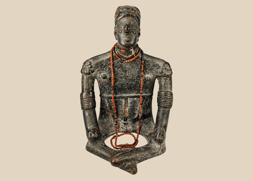 Front view of Pre-Columbian seated shamanic terracotta figure with red beads and headdress on a beige background.