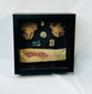 Framed display of Pre-Columbian artifacts including terracotta mask fragments, small figurines, carved ornaments, and a woven textile with red geometric pattern, set in a black shadowbox on a white background.