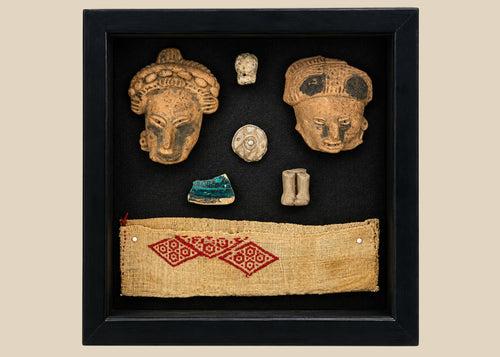 Framed display of Pre-Columbian artifacts including terracotta mask fragments, small figurines, carved ornaments, and a woven textile with red geometric pattern, set in a black shadowbox on a white background.