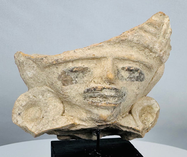 Ancient Mesoamerican terracotta face fragment featuring prominent spiral ear ornaments and traces of original pigment.