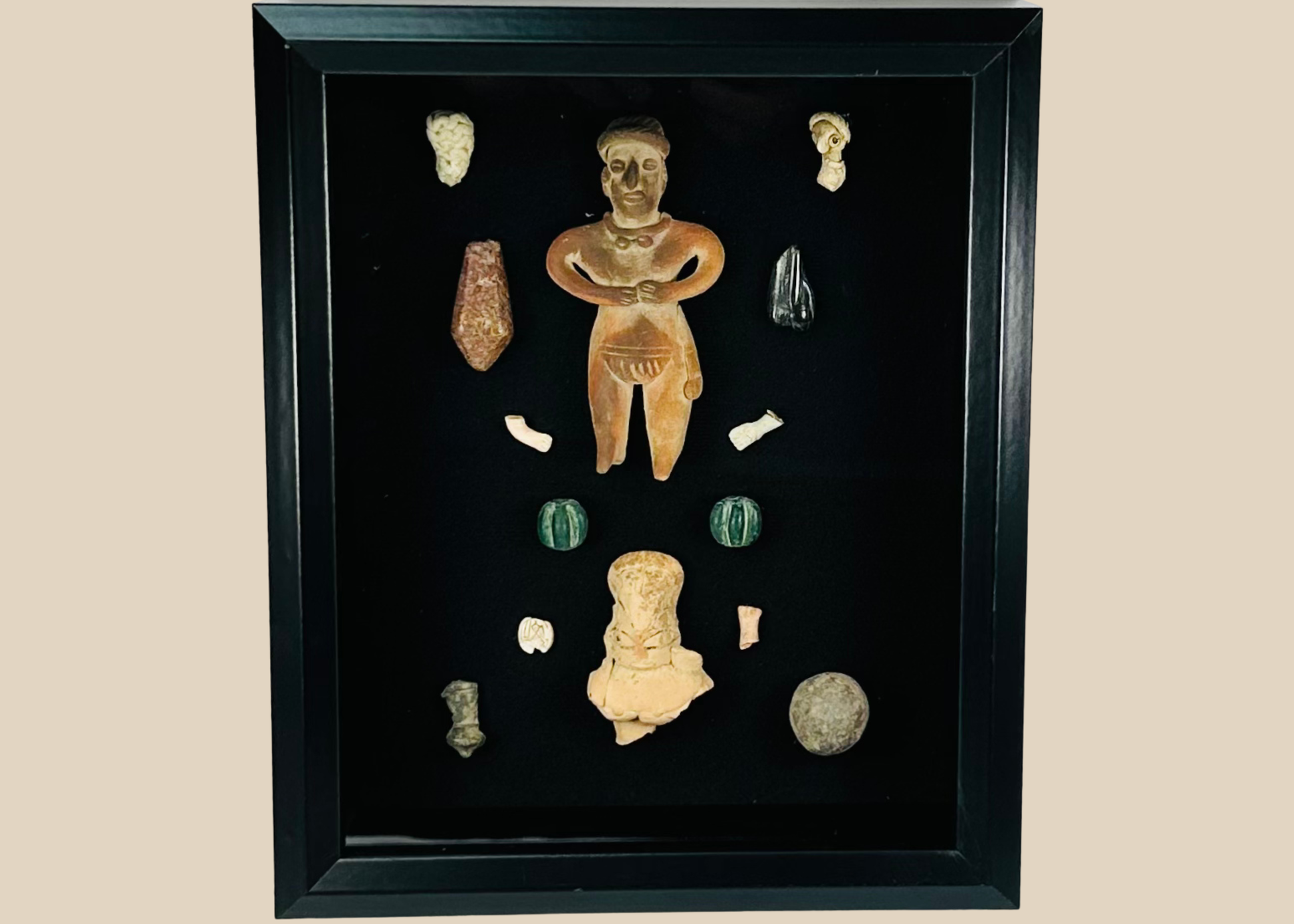 Framed Pre-Columbian artifact shadow box with terracotta, stone, and shell pieces behind glass, showing light reflections on the display surface.