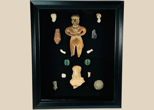 Framed Pre-Columbian artifact shadow box with terracotta, stone, and shell pieces behind glass, showing light reflections on the display surface.