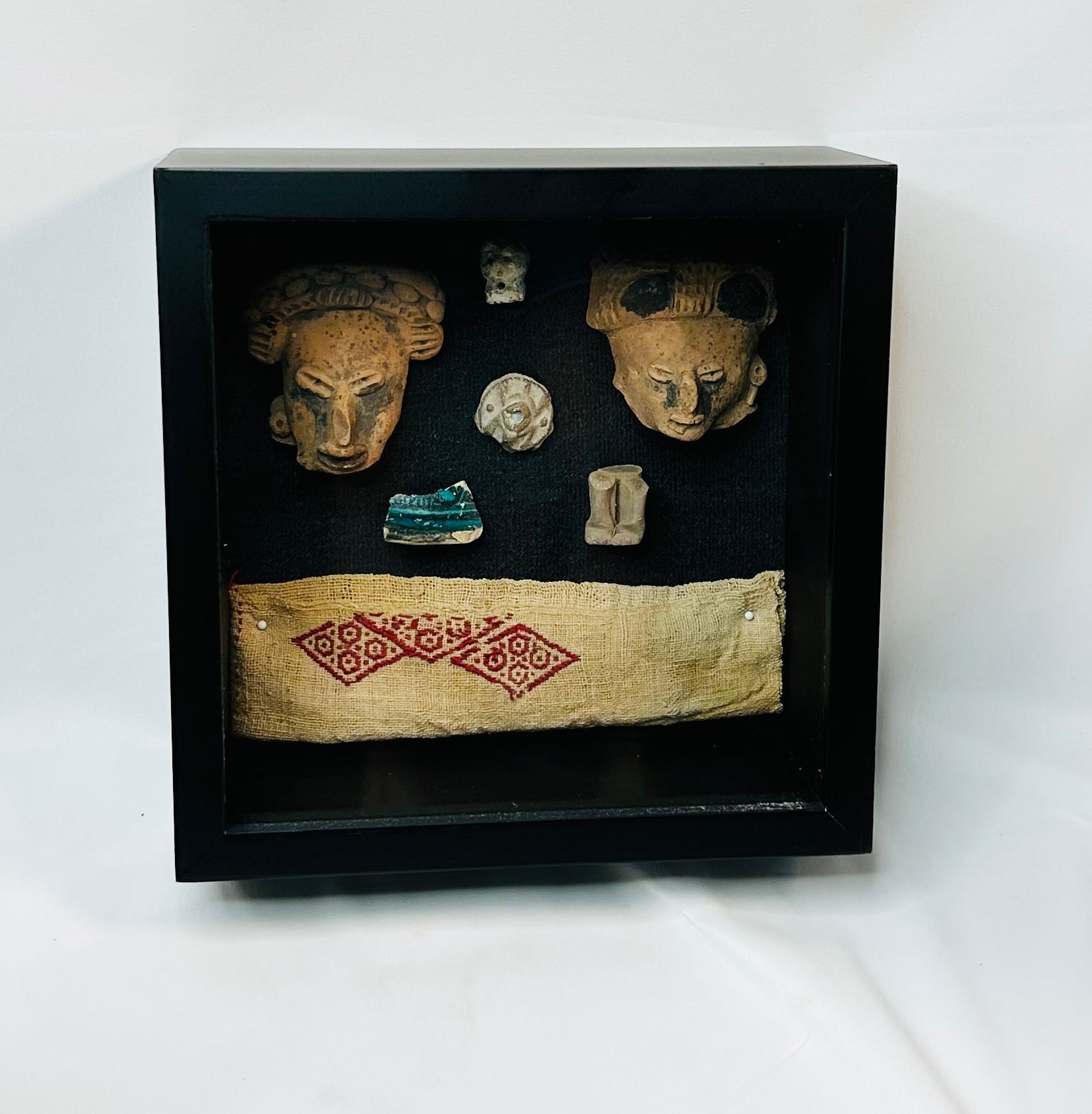 Framed display of Pre-Columbian artifacts including terracotta mask fragments, small figurines, carved ornaments, and a woven textile with red geometric pattern, set in a black shadowbox on a white background.