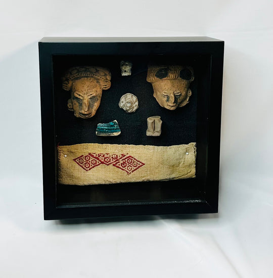 Framed display of Pre-Columbian artifacts including terracotta mask fragments, small figurines, carved ornaments, and a woven textile with red geometric pattern, set in a black shadowbox on a white background.