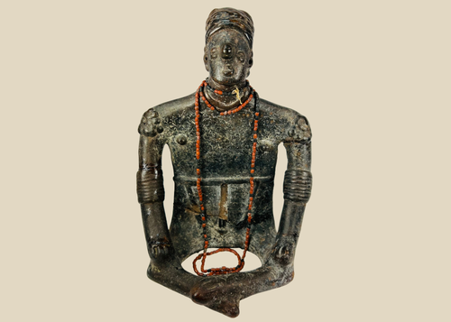 Front view of Pre-Columbian seated shamanic terracotta figure with red beads and headdress on a beige background.