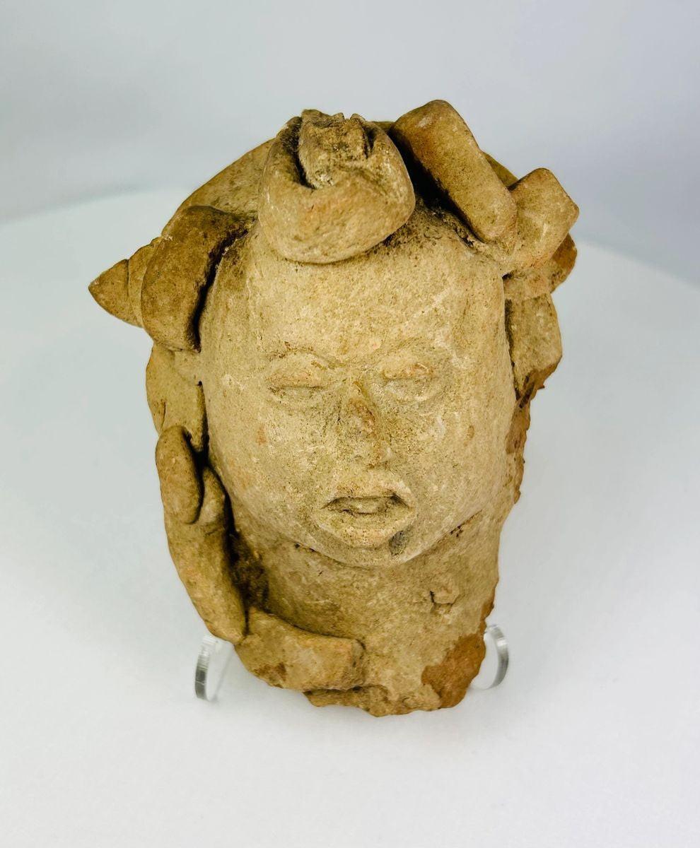 Ancient Pre-Columbian terracotta head from Mesoamerica, featuring stylized facial features with closed eyes, parted lips, and remnants of elaborate headdress elements, hand-molded from clay, dating between 1000–1500 CE, likely used as a ritual offering or funerary effigy.