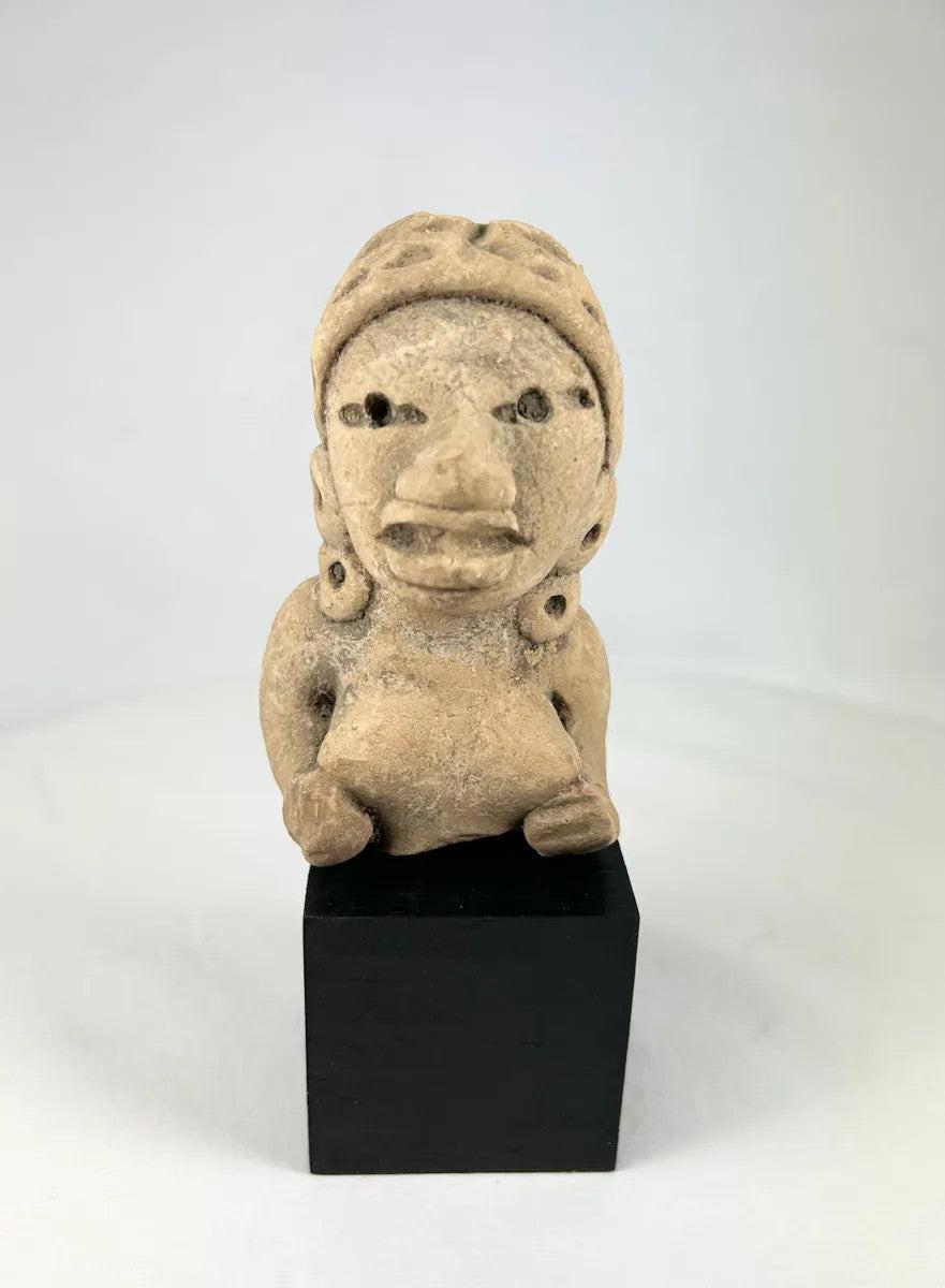 Ancient West Mexican terracotta female figurine, seated with earspools and headdress, hand-modeled with expressive facial features, mounted on a black display base, photographed on a white background.
