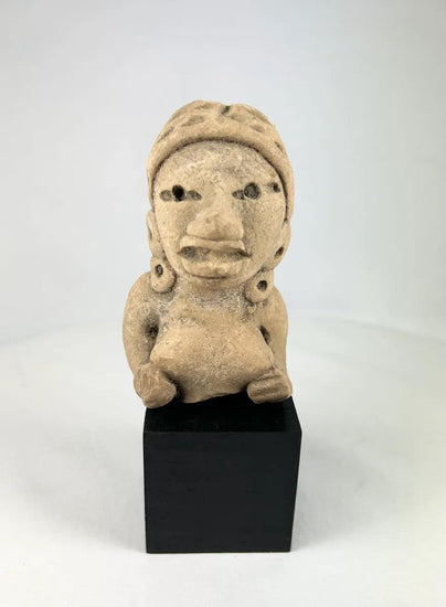 Ancient West Mexican terracotta female figurine, seated with earspools and headdress, hand-modeled with expressive facial features, mounted on a black display base, photographed on a white background.