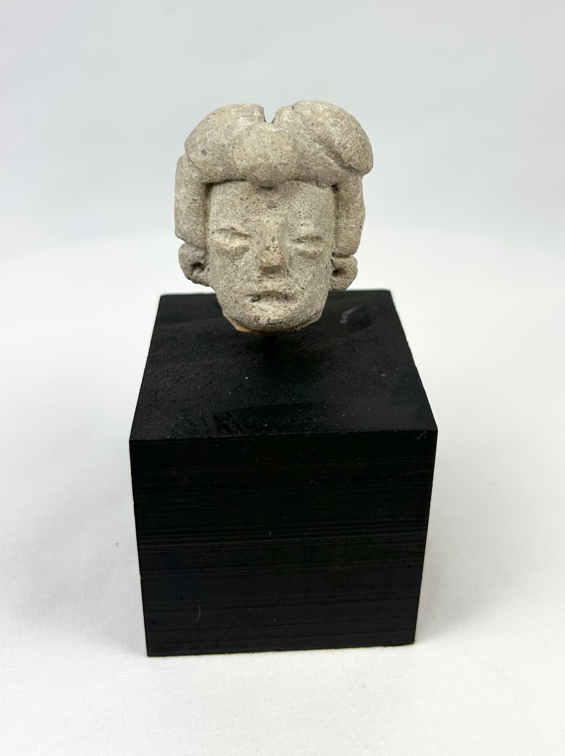 Ancient Pre-Columbian terracotta head fragment from West Mexico, Nayarit/Jalisco shaft-tomb culture, with almond-shaped eyes, straight nose, and stylized coiffure, mounted on a black display base, photographed on a white background.