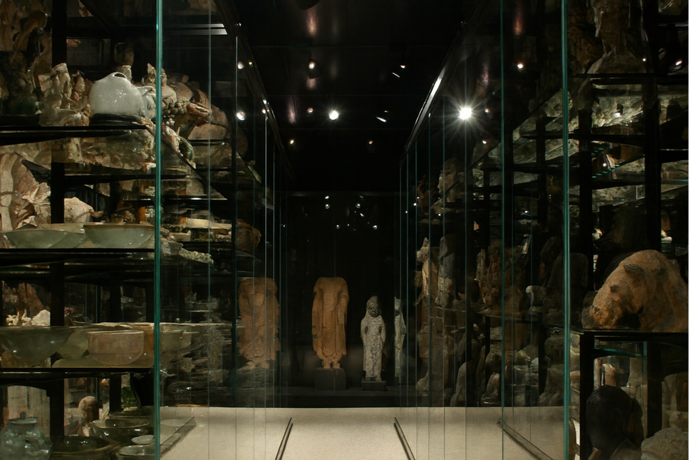 Dimly lit relic and rarity exhibit featuring ancient sculptures and relics displayed in glass cases, showcasing historical artifacts and preservation techniques.