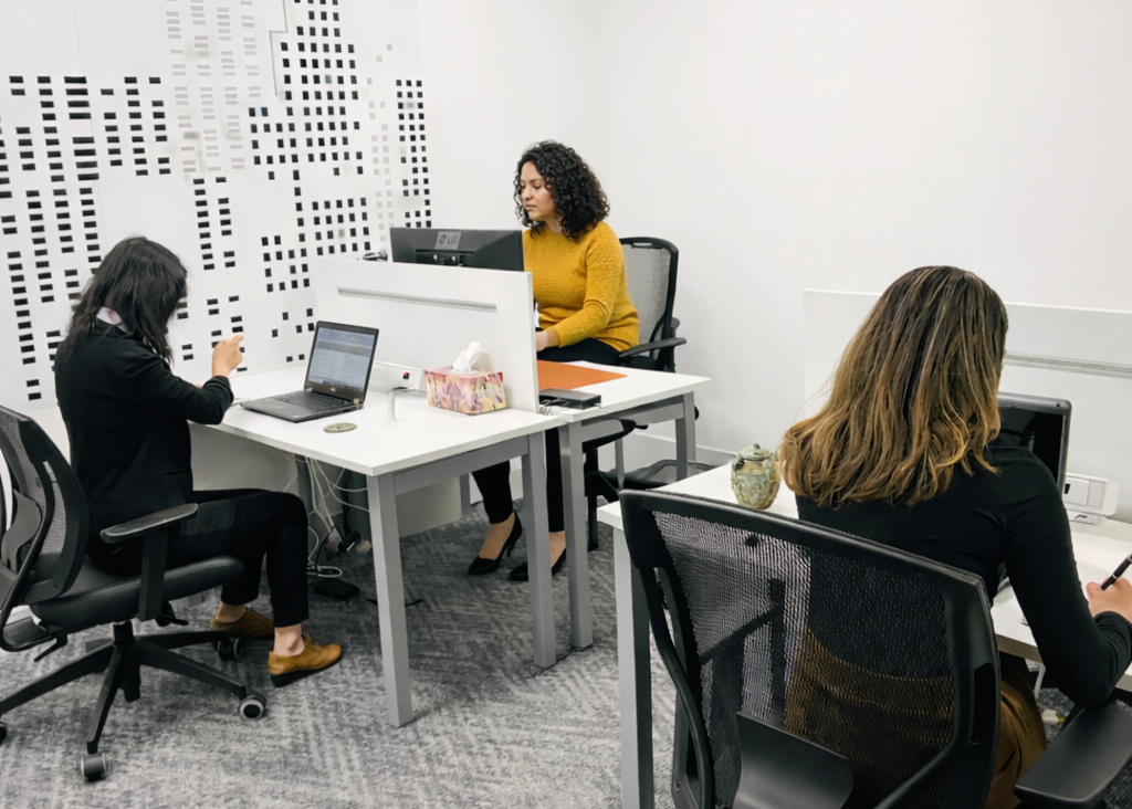 Three professional women working at laptops in a modern white office space, with a small ancient-style artifact displayed on the center desk as part of a research and cataloging process.