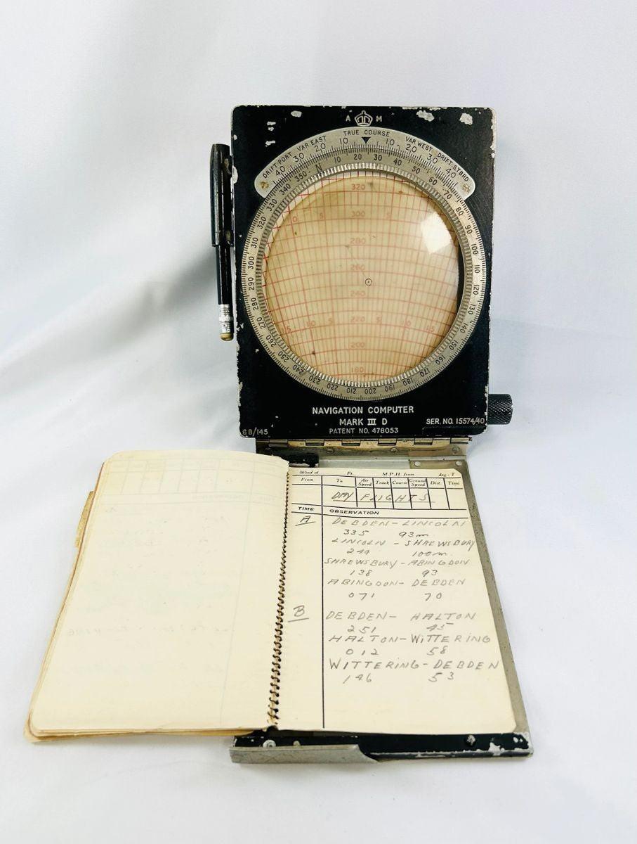 RAF WWII navigational computer with rotating aluminum dial, Bakelite adjustments, original fabric straps, pencil, and open flight notebook showing handwritten navigator’s notes.