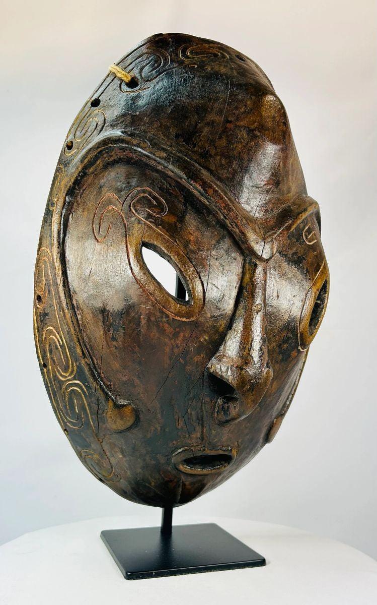 Early 20th-century Sepik River tribal ceremonial mask, hand-carved from wood with elongated oval form, large almond-shaped eyes, incised swirling patterns, and traces of natural pigments, mounted on a display stand; traditional artifact used in ancestral rituals and cultural ceremonies of Papua New Guinea.