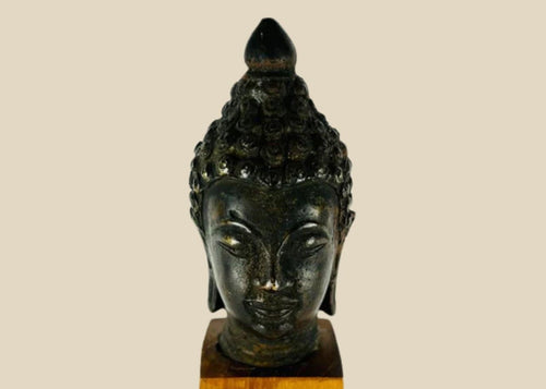 Antique Southeast Asian cast bronze Buddha head, likely from Thailand or Burma, 18th–19th Century CE. Features serene facial expression, closed eyes, gentle smile, ushnisha symbolizing wisdom, and intricately modeled hair curls. Dark natural patina with wooden base, reflecting Theravāda Buddhist devotional artistry and meditation iconography.