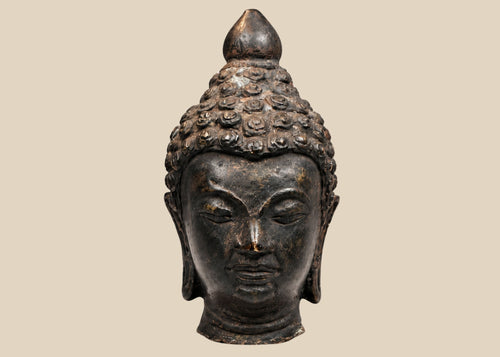 Antique Southeast Asian cast bronze Buddha head, likely from Thailand or Burma, 18th–19th Century CE. Features serene facial expression, closed eyes, gentle smile, ushnisha symbolizing wisdom, and intricately modeled hair curls. Dark natural patina with wooden base, reflecting Theravāda Buddhist devotional artistry and meditation iconography.