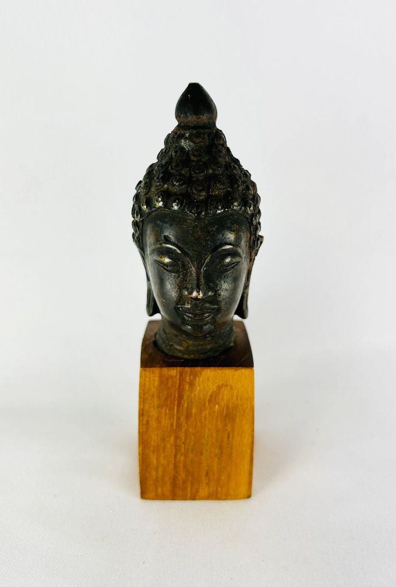 Antique Southeast Asian cast bronze Buddha head, likely from Thailand or Burma, 18th–19th Century CE. Features serene facial expression, closed eyes, gentle smile, ushnisha symbolizing wisdom, and intricately modeled hair curls. Dark natural patina with wooden base, reflecting Theravāda Buddhist devotional artistry and meditation iconography.