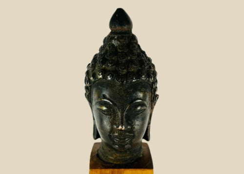 Antique Southeast Asian cast bronze Buddha head, likely from Thailand or Burma, 18th–19th Century CE. Features serene facial expression, closed eyes, gentle smile, ushnisha symbolizing wisdom, and intricately modeled hair curls. Dark natural patina with wooden base, reflecting Theravāda Buddhist devotional artistry and meditation iconography.