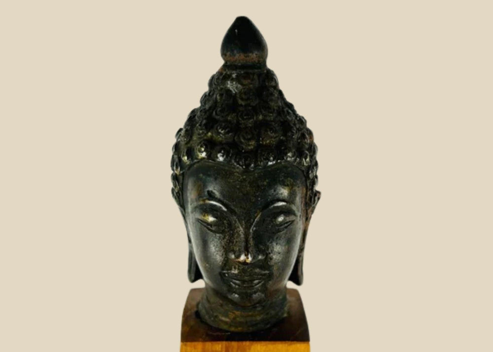 Antique Southeast Asian cast bronze Buddha head, likely from Thailand or Burma, 18th–19th Century CE. Features serene facial expression, closed eyes, gentle smile, ushnisha symbolizing wisdom, and intricately modeled hair curls. Dark natural patina with wooden base, reflecting Theravāda Buddhist devotional artistry and meditation iconography.