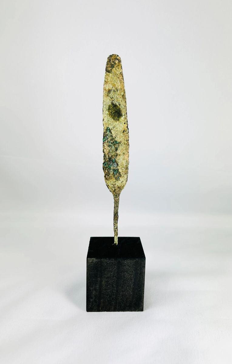 Ancient bronze spearhead from the Early to Late Bronze Age (circa 3300–1200 BCE). Leaf-shaped blade with tapered point, mounted on a modern display base. Exhibits natural patina and surface oxidation, reflecting early metallurgical craftsmanship and use in hunting or combat.