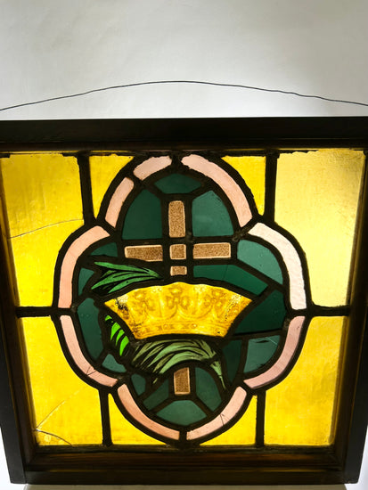 Early 20th-century stained glass panel featuring a central cross and crown motif in amber, green, and pink glass set in a wooden frame, approximately 16 by 16 inches.