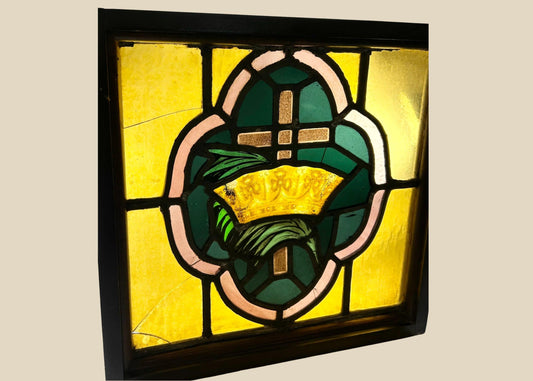 Early 20th-century stained glass panel featuring a central cross and crown motif in amber, green, and pink glass set in a wooden frame, approximately 16 by 16 inches.