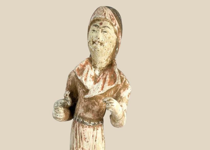 Tang Dynasty low-fired terracotta figure of a female court attendant, standing with hands clasped, wearing flowing robes with traces of red and white pigments, serene expression, Tang hairstyle, 13 inches tall, surface mineral deposits and patina, circa 618–907 CE.