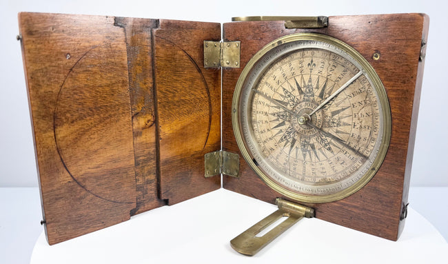 Front-facing open walnut surveyor’s compass with silvered circular dial, central magnetic needle, and hinged wooden lid.