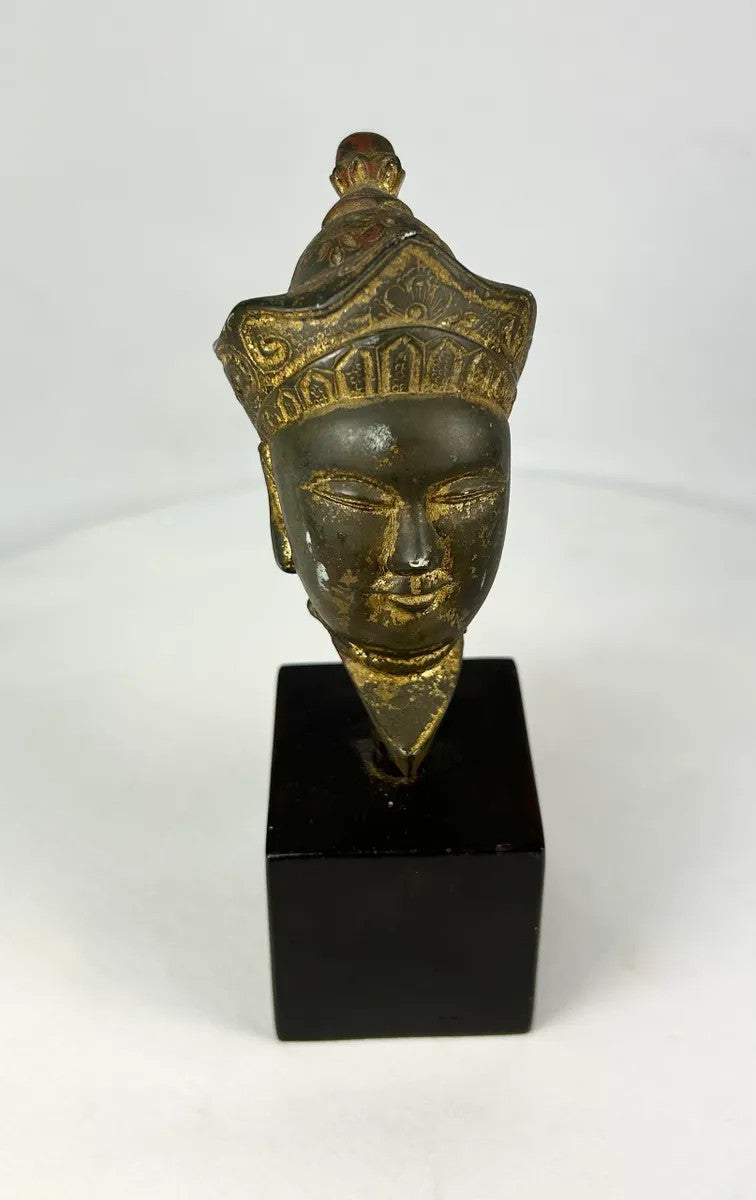 Antique Tibetan gilded bronze Buddha head with ornate crown, serene facial features, and traces of gilding, mounted on a black display base, photographed on a white background.