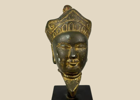 Antique Tibetan gilded bronze Buddha head with ornate crown, serene facial features, and traces of gilding, mounted on a black display base, photographed on a white background.