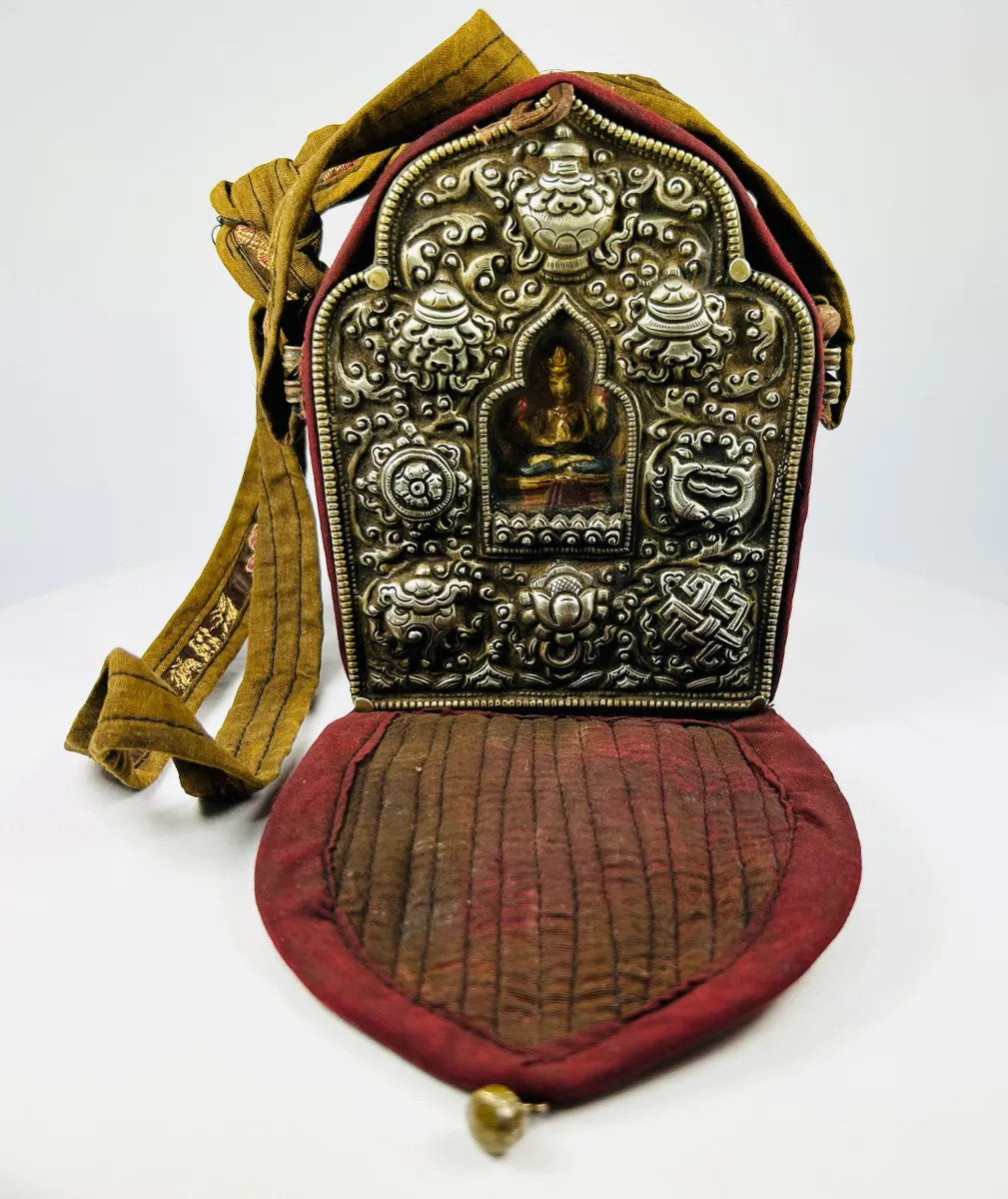 19th-century Tibetan portable shrine (gau) with silver repoussé Buddhist motifs and central glazed Buddha figure, set in embroidered red and gold fabric case with strap, photographed on a white background.